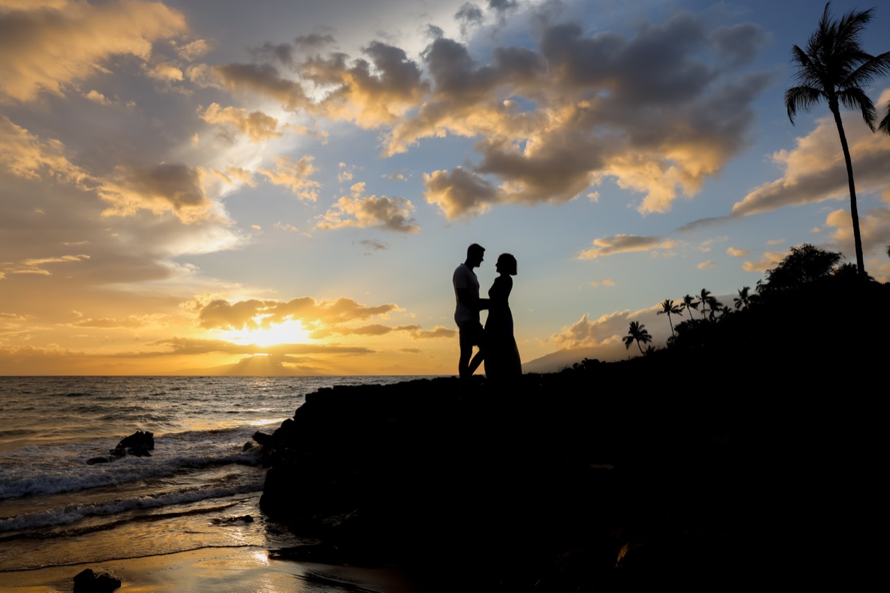 A silhouetted couple stands above the ocean beneath golden sunset clouds.